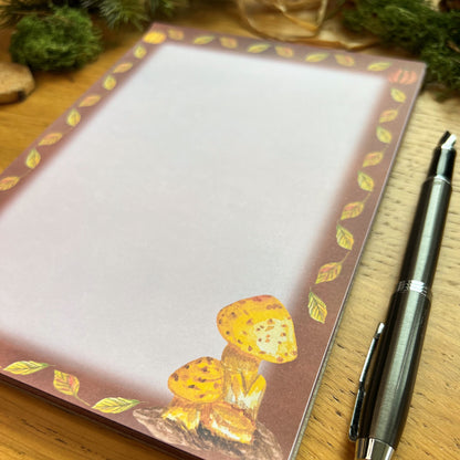 Autumnal mushroom illustrated notepad with warm brown leaf and pumpkin illustrated border around the page. The writing paper is blank. Notepad is on a wooden desk decorated with green moss, straw, pine cones and wooden slices and a fountain ink pen