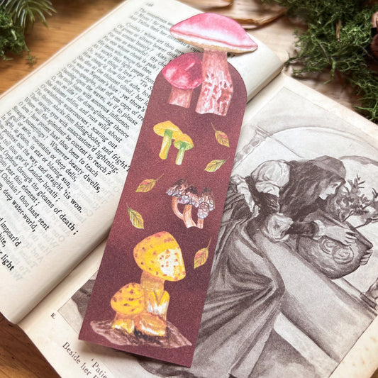 Fall Forage bookmark fully illustrated with mushrooms and leaves with a deep warm brown background, with cut out of mushroom on the top. Bookmark resting on an open poetry book sat on a wooden desk decorated with green moss and straw