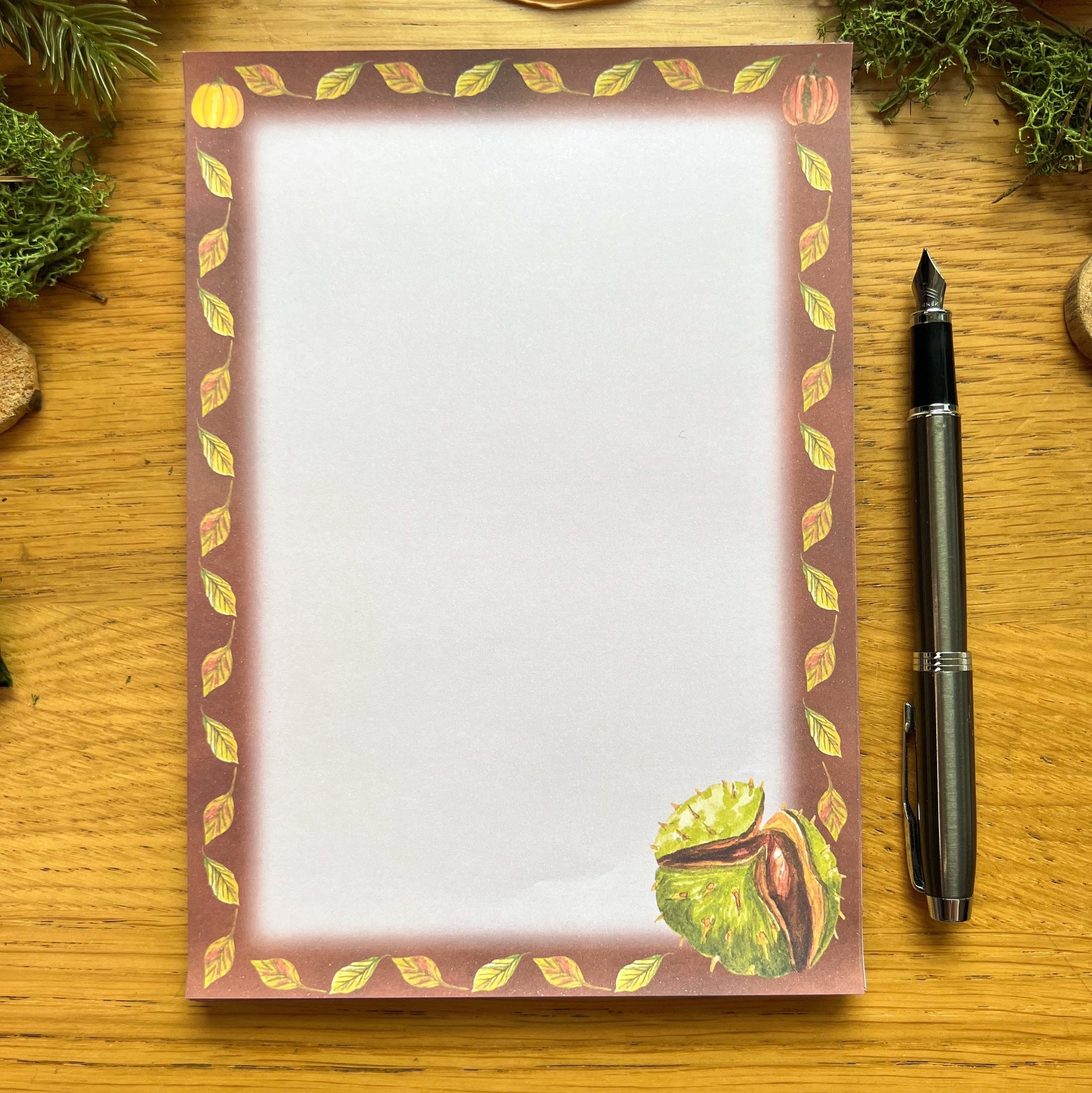 Autumnal conker illustrated notepad with warm brown leaf and pumpkin illustrated border around the page. The writing paper is blank. Notepad is on a wooden desk decorated with green moss, straw, pine cones and wooden slices and a fountain ink pen