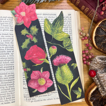 2 winters bloom bookmarks one decorated with illustrated rose, hellebore, holly and poinsettia, the other with thistle and ivy, both sat in the open pages of a book