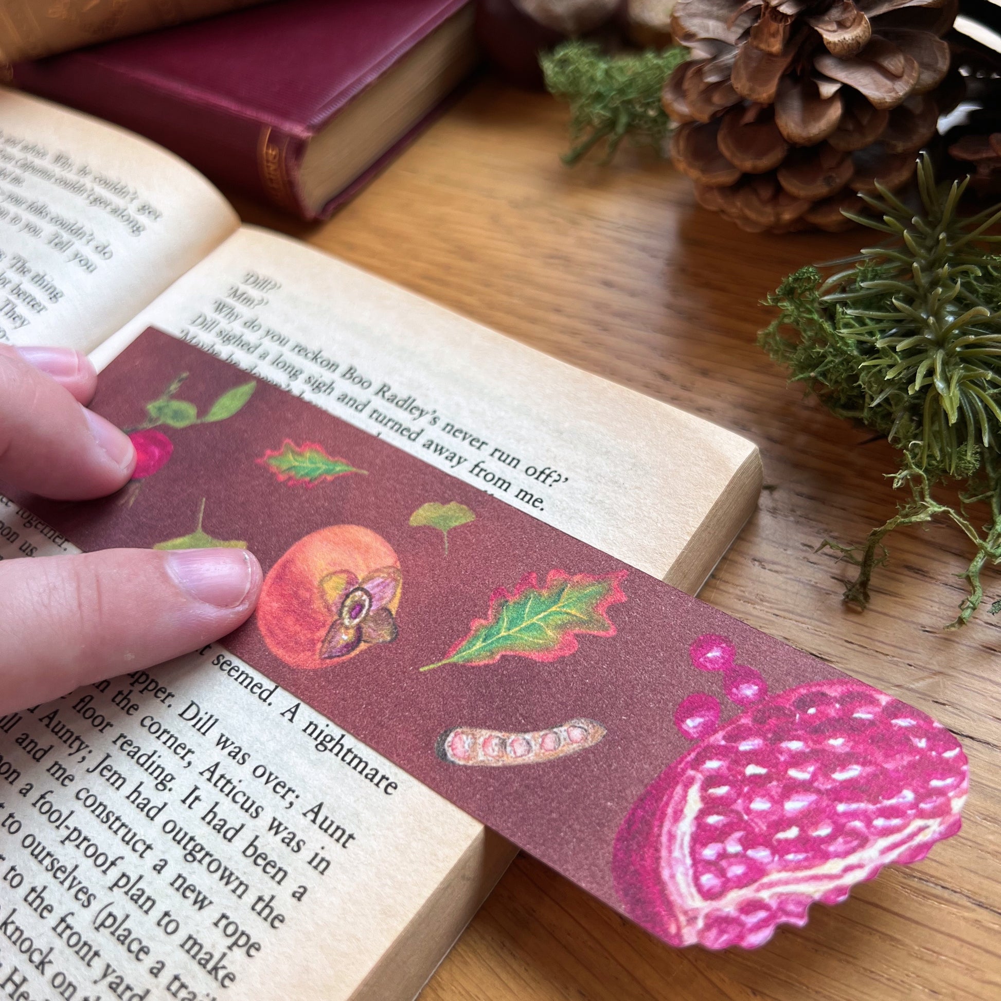 Woodland Walk Fruits Bookmark being used to read the open pages of a book. Bookmark features a pomegranate cut out at the top, and has autumnal leaves, persimmon, rose hips and seed pods illustrated across a warm toned brown background.