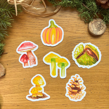 Fall Forage Die Cut Easy Peel Stickers laid out on a wooden desk decorated with green moss, straw, pines and wooden slices. Stickers include pumpkins, mushrooms, conker and pine cone