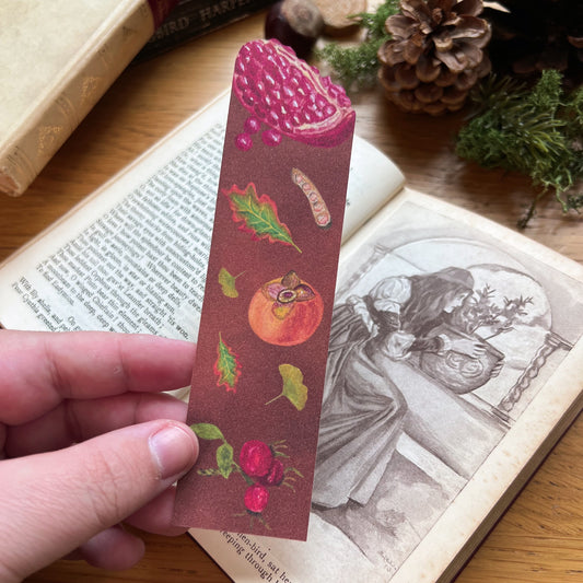 Woodland Walk Fruits Bookmark held in front of an open book. Bookmark features a pomegranate cut out at the top, and has autumnal leaves, persimmon, rose hips and seed pods illustrated across a warm toned brown background.