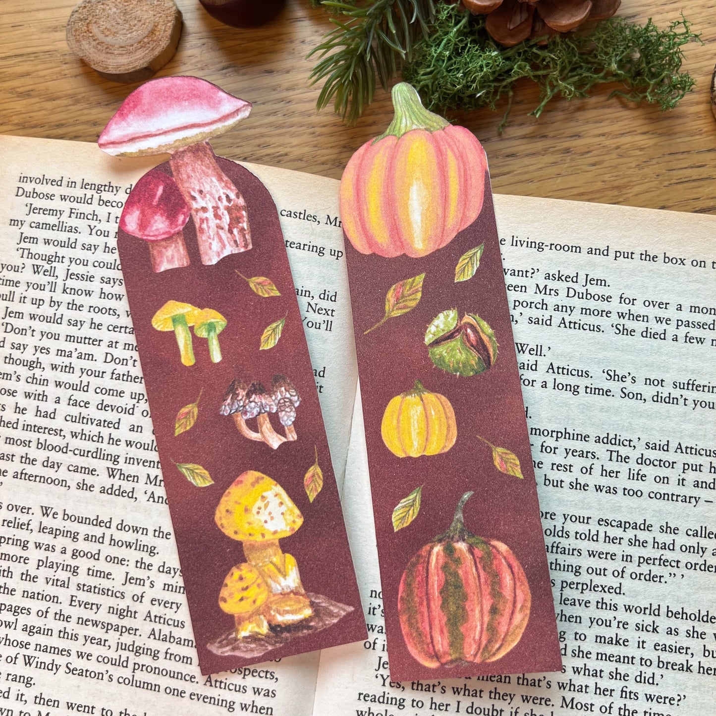 Fall Forage Pumpkin Bookmark featuring two orange and one yellow pumpkins with autmnal leabes tumbling dow the illustrated bookmark. An orange Cinderella pumpkin is cut out at the top, and the bookmark is resting on the open pages of a book next to the Fall Forage Mushrooms bookmark which features Mushrooms acorss the warm tones instead.