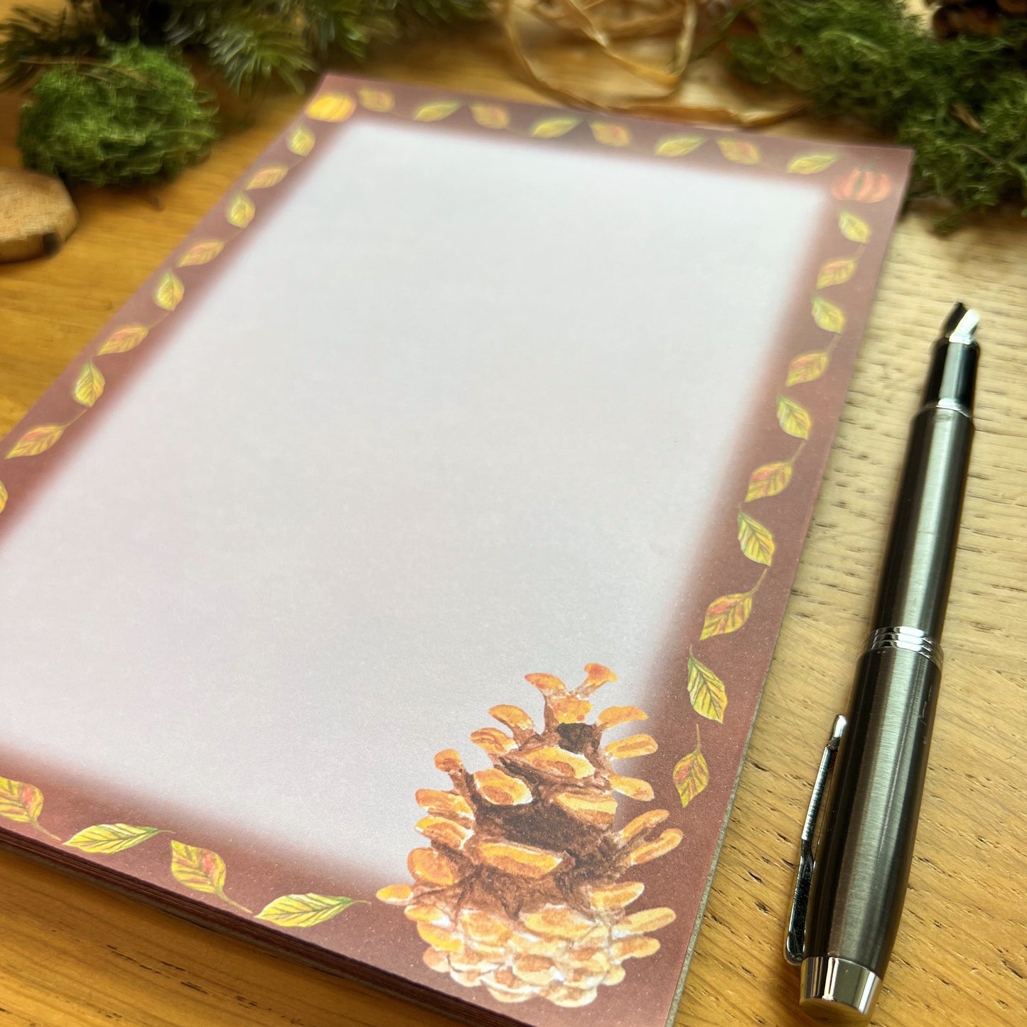 Autumnal pine cone illustrated notepad with warm brown leaf and pumpkin illustrated border around the page. The writing paper is blank. Notepad is on a wooden desk decorated with green moss, straw, pine cones and wooden slices and a fountain ink pen