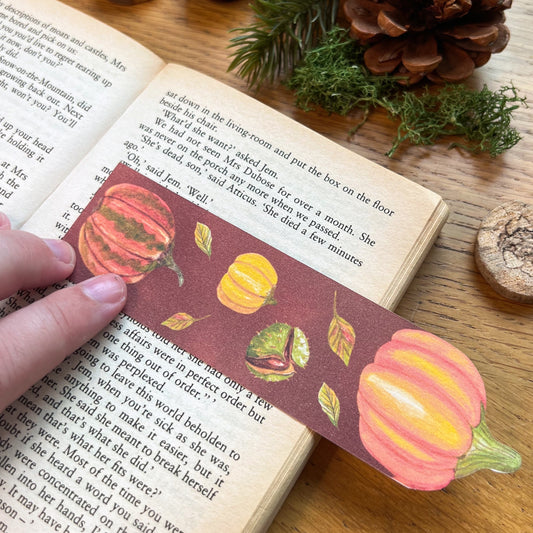 Fall Forage Pumpkin Bookmark featuring two orange and one yellow pumpkins with autmnal leabes tumbling dow the illustrated bookmark. An orange Cinderella pumpkin is cut out at the top, and the bookmark is being used to read the open pages of a book