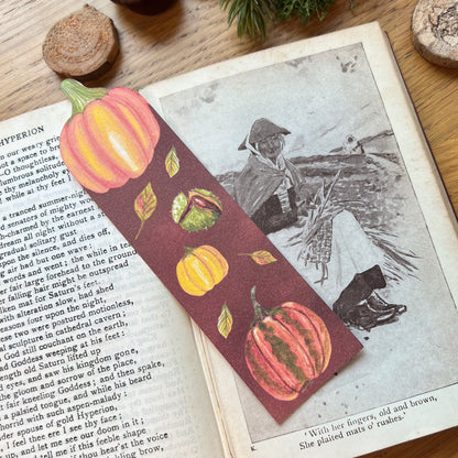 Fall Forage Pumpkin Bookmark featuring two orange and one yellow pumpkins with autmnal leabes tumbling dow the illustrated bookmark. An orange Cinderella pumpkin is cut out at the top, and the bookmark is resting on the open pages of a book with an ink illustration of woman resting whilst working in the fields