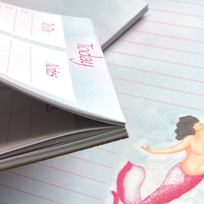 Close up of Blossom planner pad from the side bending pages, in front of stack of notepads. Blossom mermaid is shown close up on a lined pad, has a light and magenta pink tail shaped like petals, with Caucasian skin tone, subtle top surgery scars and mid length black hair flowing.