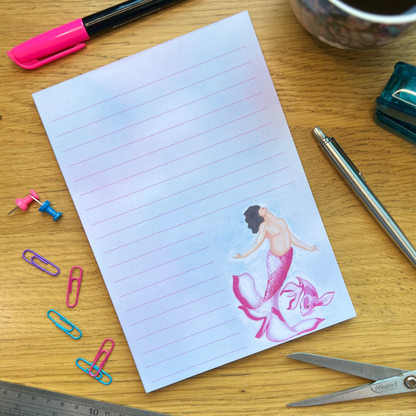 Blossom lined notepad washed with light blue and pink ancross the page, illustrated with Blossom mermaid and koi bottom right. Blossom has a light and magenta pink tail shaped like petals, with Caucasian skin tone, subtle top surgery scars and mid length black hair flowing. A matching colours pink koi swims next to them. Notepad is on a wooden desk surrounded by pens and stationery items to decorate.