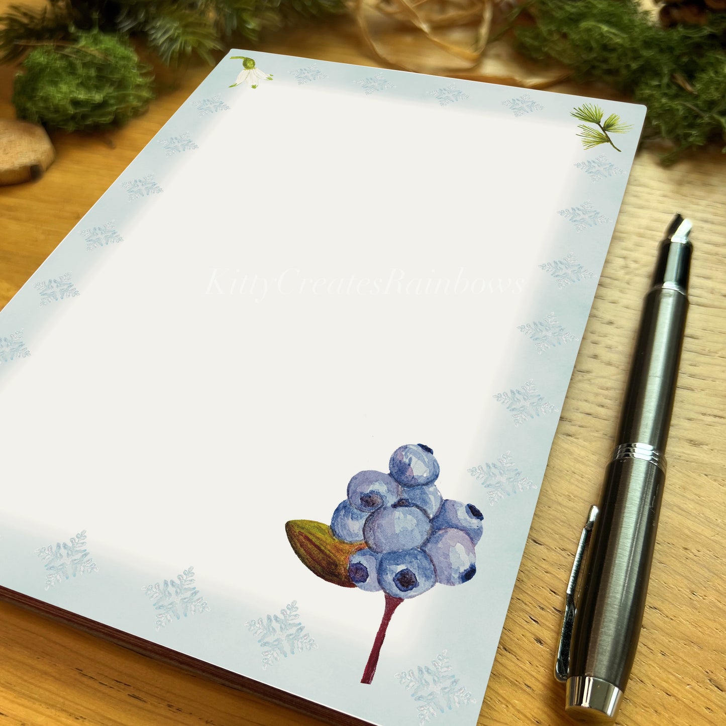 Northern blueberry illustrated notepad page with icy blue border on a wooden desk with a fountain pen