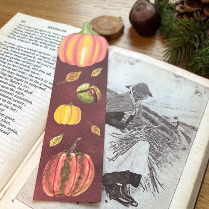Fall Forage Pumpkin Bookmark featuring two orange and one yellow pumpkins with autmnal leabes tumbling dow the illustrated bookmark. An orange Cinderella pumpkin is cut out at the top, and the bookmark is resting on the open pages of a book