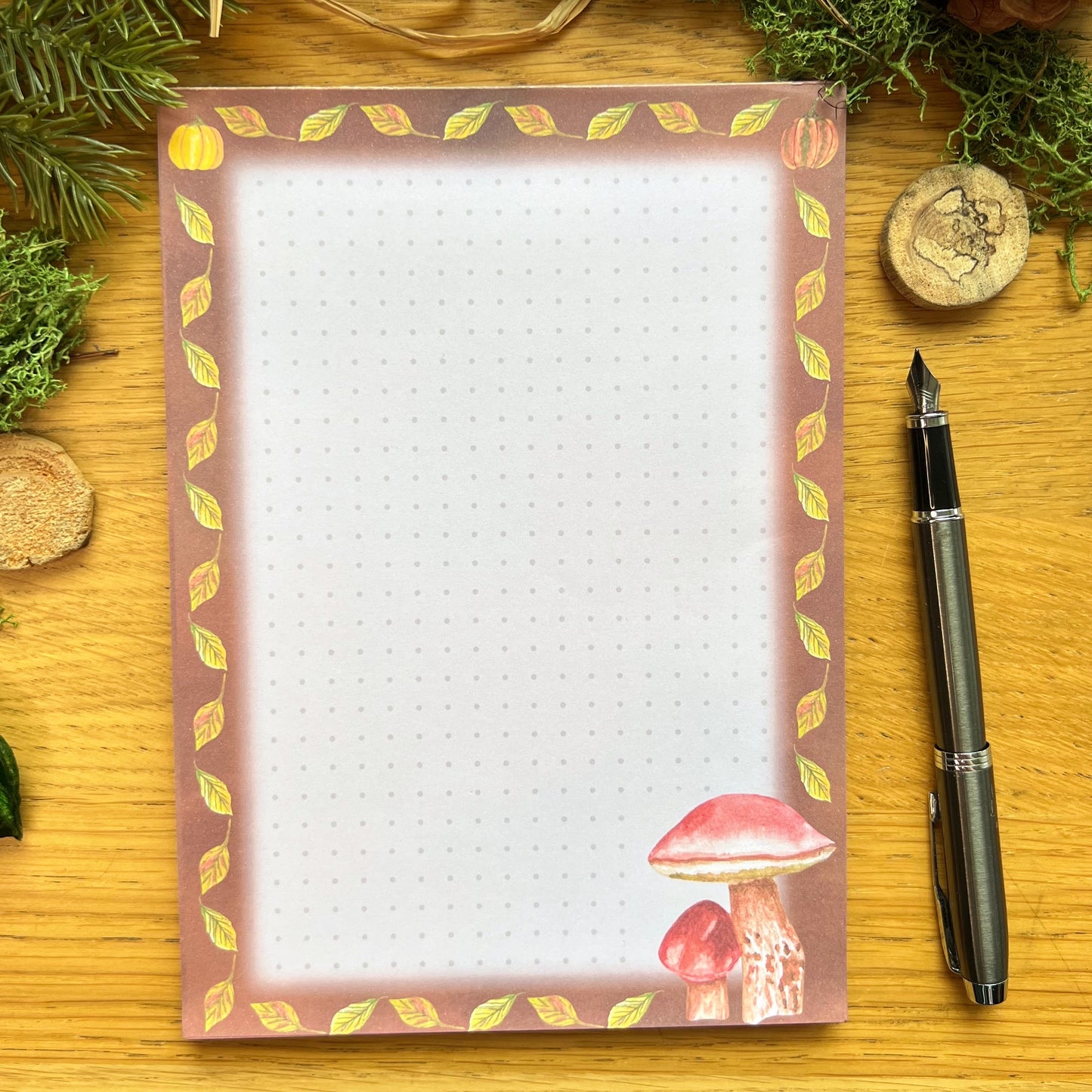 Autumnal mushroom illustrated notepad with warm brown leaf and pumpkin illustrated border around the page. The writing paper is subtly dotted in a grid. Notepad is on a wooden desk decorated with green moss, straw, pine cones and wooden slices and a fountain ink pen