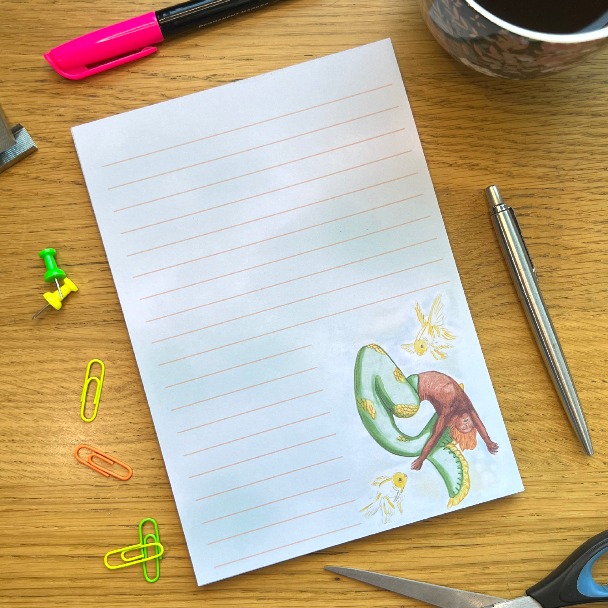 Citrus lined notepad sat in a wooden desk decorated and surrounded by pens and other stationery items. Notepad is washed with green and blue and features Citrus bottom right. Citrus mermaid dancing in a whirlwind shape bending backwards accompanied by two white and yellow goldfiish. Citrus has orange toned skin and a green tail with patches of yellow scales.