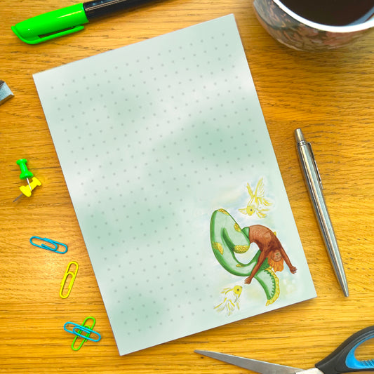 Citrus dotted notepad sat in a wooden desk decorated and surrounded by pens and other stationery items. Notepad is washed with green and blue and features Citrus bottom right. Citrus mermaid dancing in a whirlwind shape bending backwards accompanied by two white and yellow goldfiish. Citrus has orange toned skin and a green tail with patches of yellow scales.
