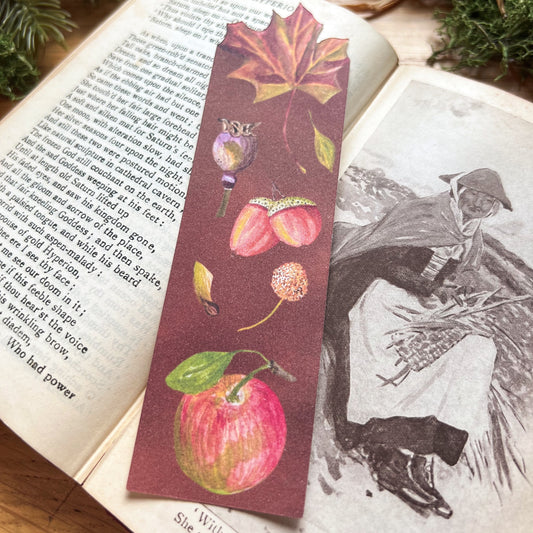 Woodland Walk bookmark fully illustrated with seed pods, acorn, apple and leaves with a deep warm brown background, with cut out of maple leaf on the top. Bookmark resting on an open poetry book sat on a wooden desk decorated with green moss and straw