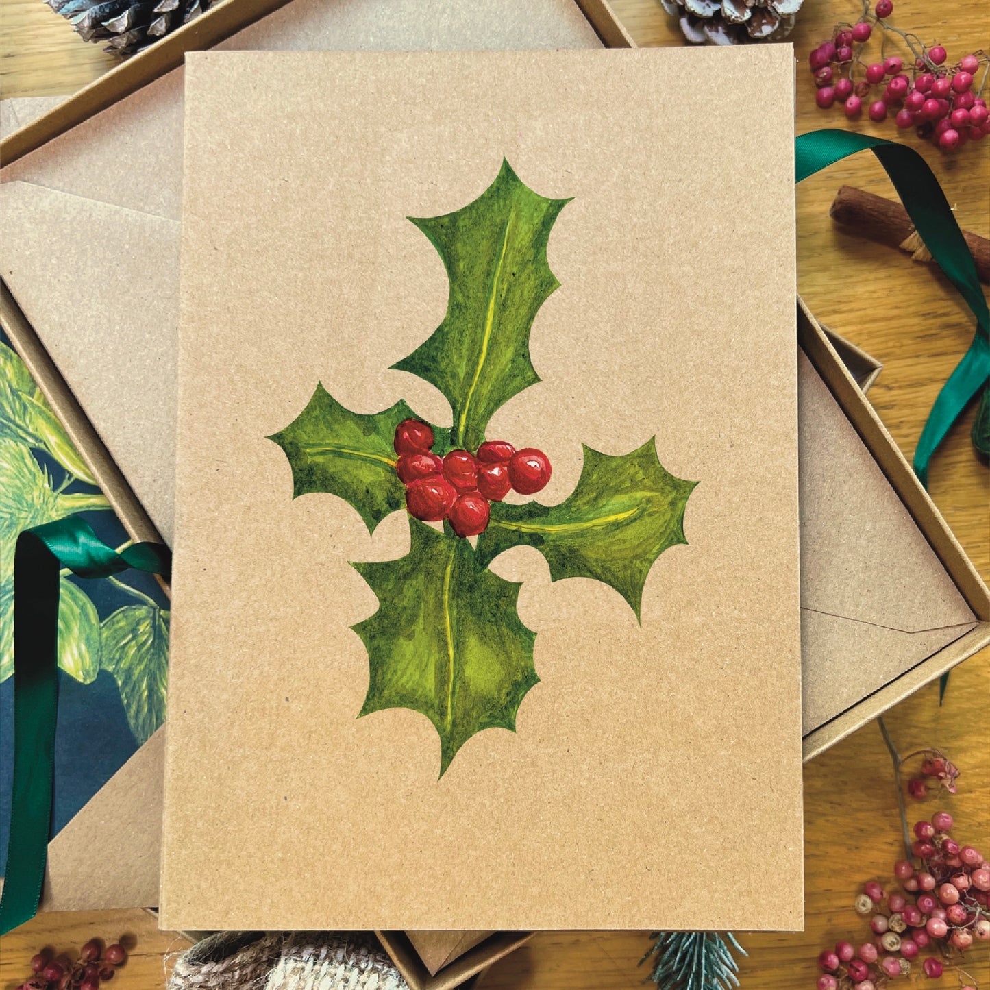 English Holly sprig with berries illustrated in watercolour attached to brown Manila recycled greetings card on a wooden desk