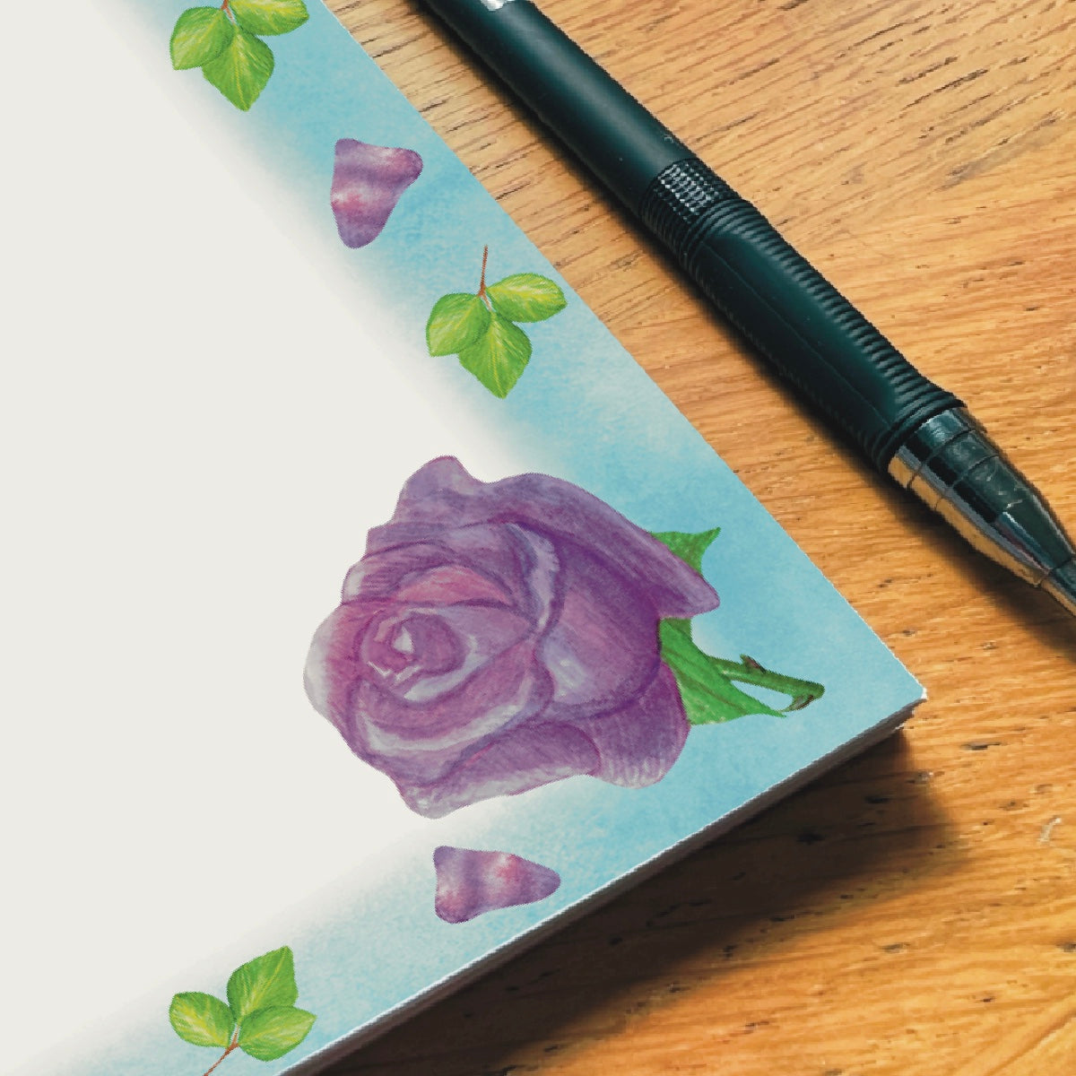 Close up corner of lavender rose illustrated blank notepad with sky blue border decorated with lavender petals and leaves
