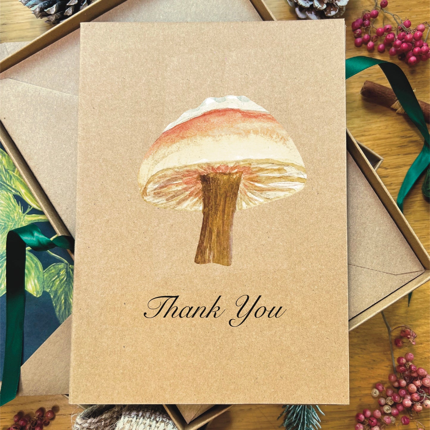 Brown manila recycled greetings card featuring a watercolour painted snow topped Milkcap mushroom. Text underneath the mushroom says Thank You printed in italic black script.