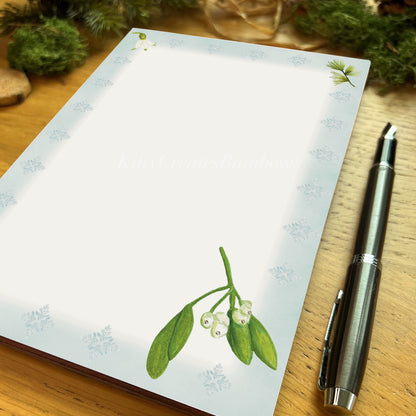 Mistletoe spit with white berries illustrated notepad page with icy blue border on a wooden desk with a fountain pen