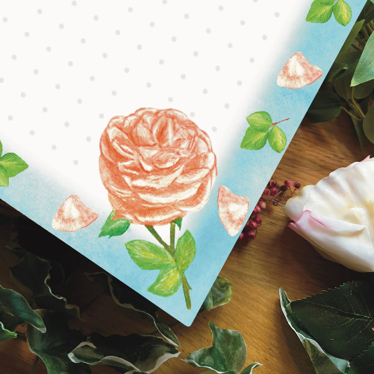 Close up corner of peach rose illustrated dotted notepad with sky blue border decorated with peach petals and leaves