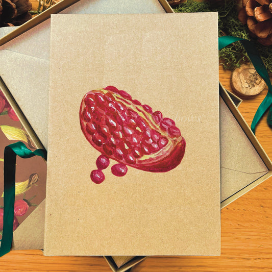 Red pomegranate slice watercolour illustrated recycled Manila blank greetings card laying on a box next to accompanying illustrated inlay envelope