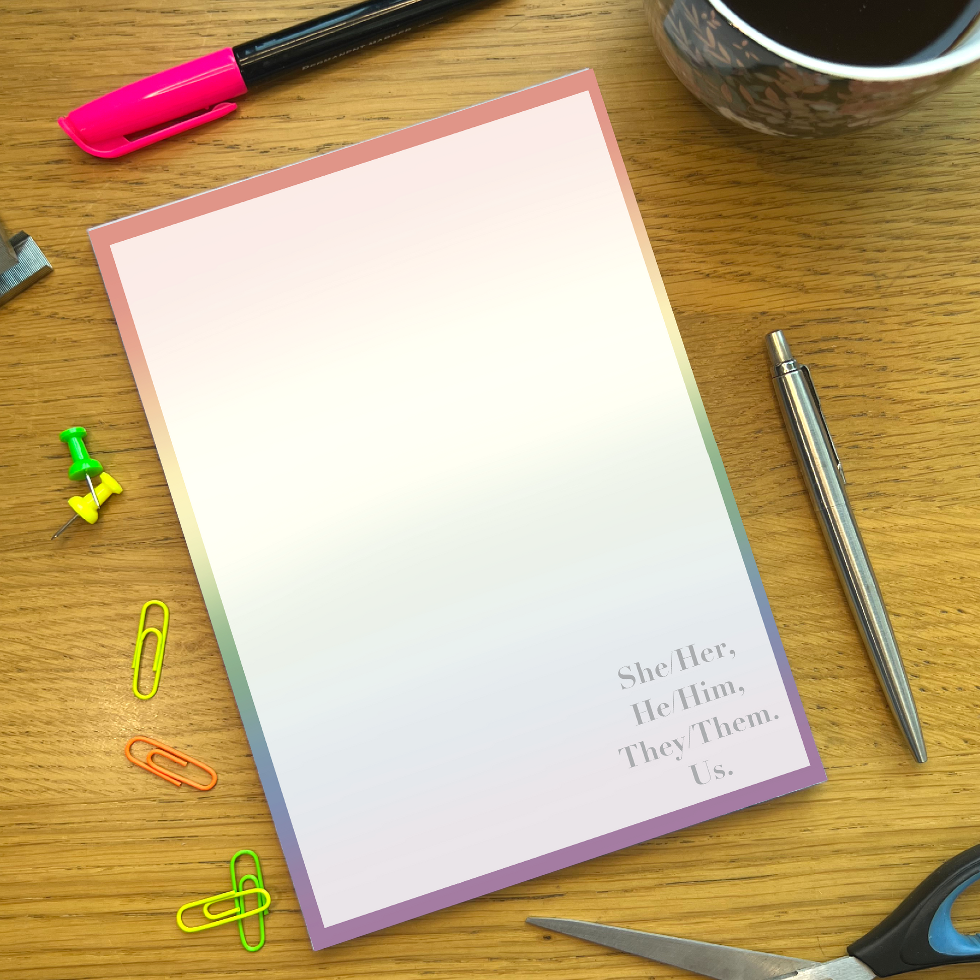 Rainbow pride notepad with quote She/her, He/him, They/them, Us, on a wooden desk with stationery