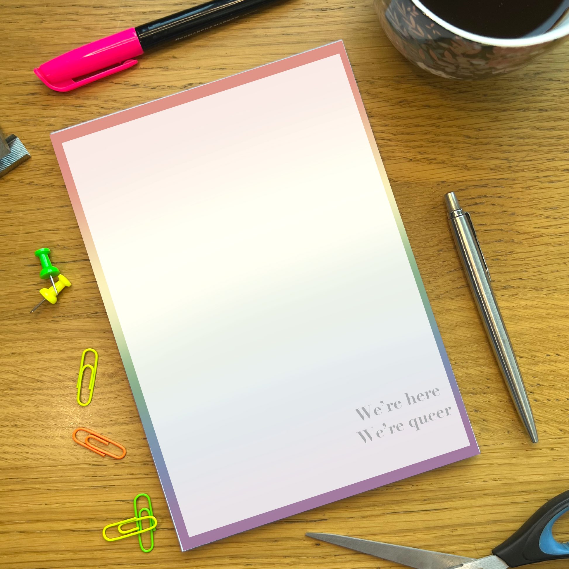Rainbow pride notepad with quote We’re Here, We’re Queer, on a wooden desk with stationery