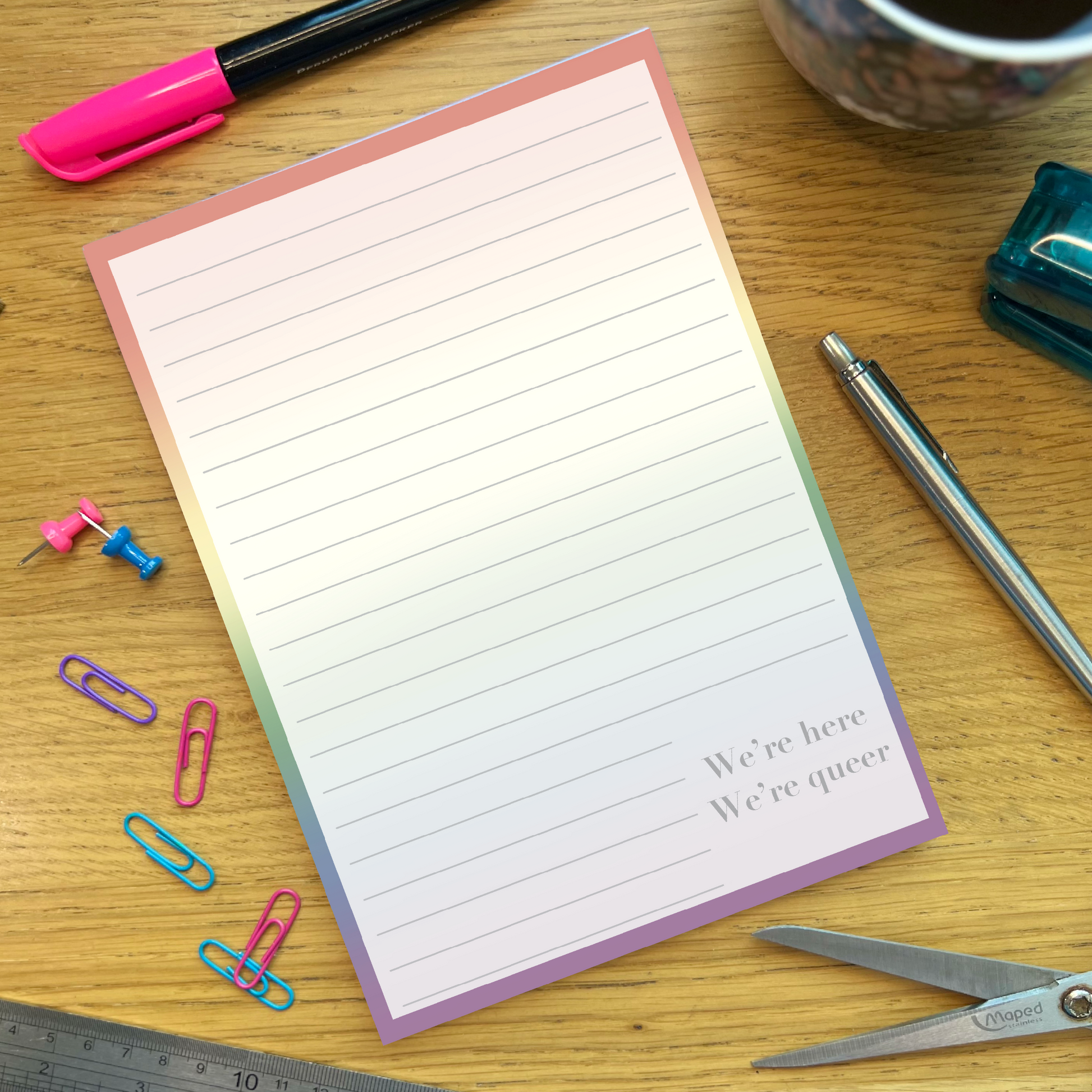 Rainbow pride lined notepad with quote We’re Here, We’re Queer, on a wooden desk with stationery