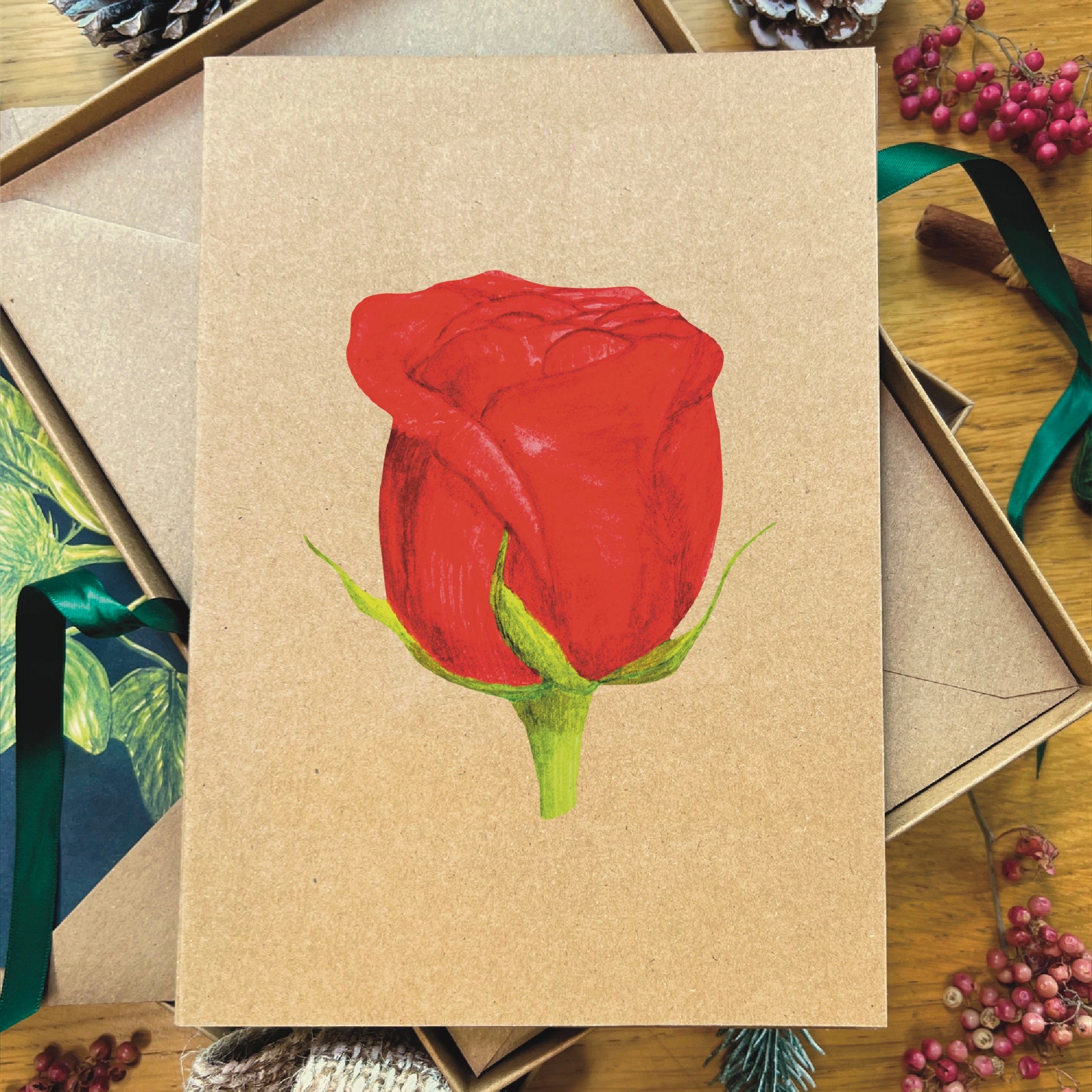 Red hybrid tea rose illustrated in watercolour attached to brown Manila recycled greetings card on a wooden desk