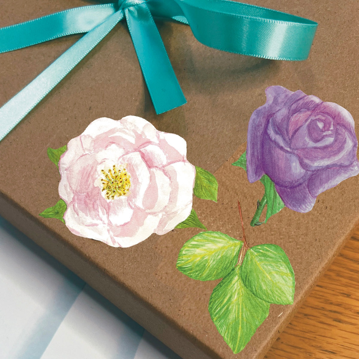 A blossoming pink and lavender rose set of die cut stickers with and easier to peel edge on a gift box wrapped with a blue ribbon