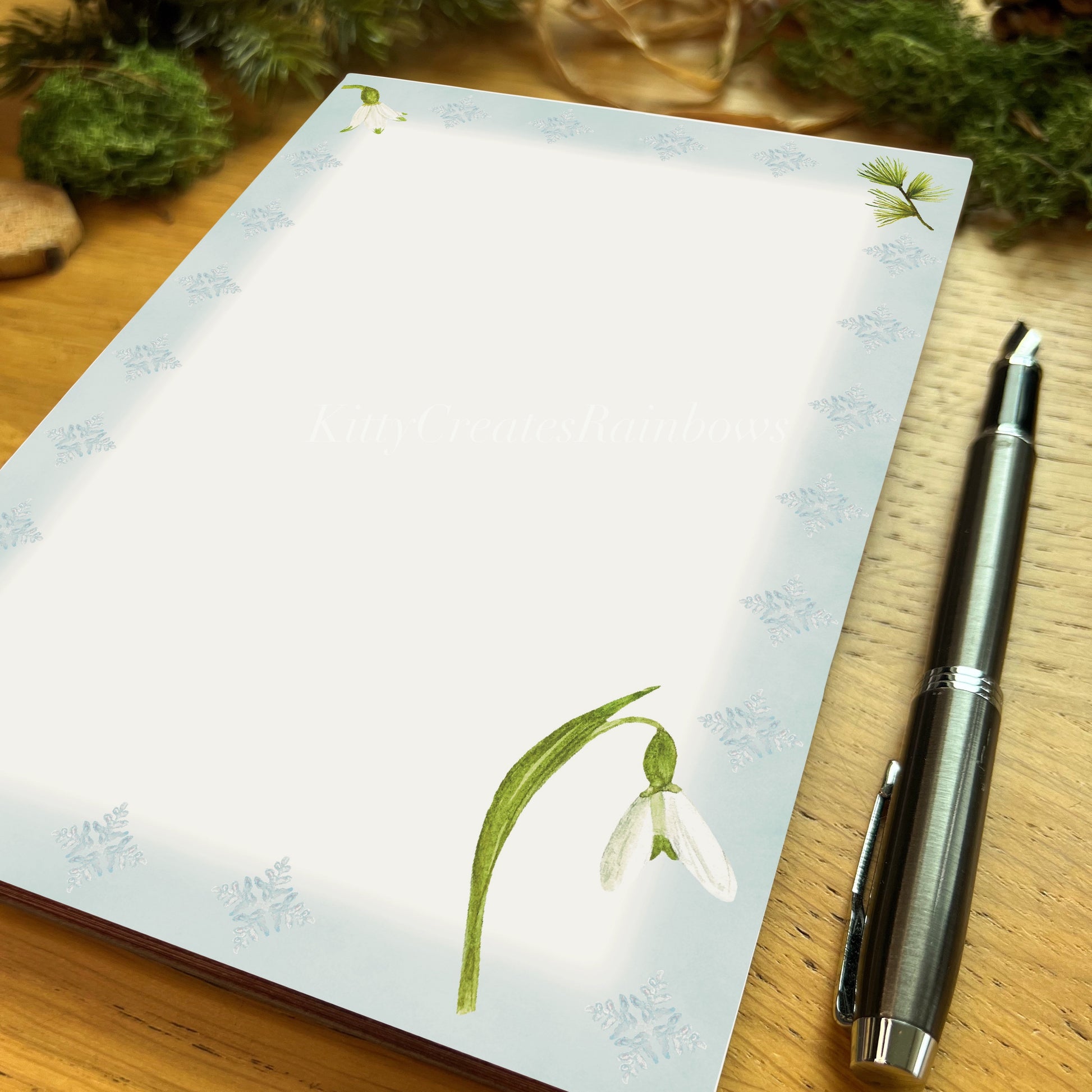 Snowdrop illustrated notepad page with icy blue border on a wooden desk with a fountain pen