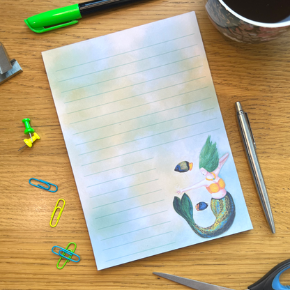 Stripe lined notepad on a wooden desk surrounded by stationery.  Stripe has a blue green and gold striped tail, green flowing long hair and is pointing towards a glowing yellow light, and two fish swim nearby.
