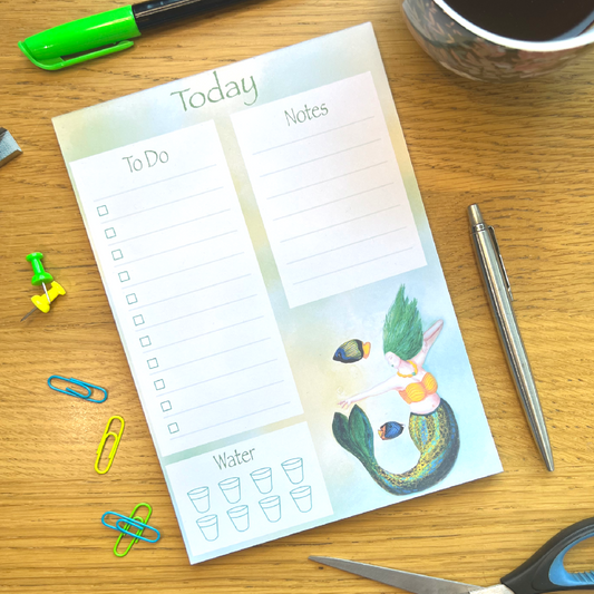 Stripe Planner pad on a wooden desk surrounded by stationery. Pad features 3 white boxes, To Do on left, Note on right, Water bottom left, and illustrated with Stripe bottom right.  Stripe has a blue green and gold striped tail, green flowing long hair and is pointing towards a glowing yellow light, and two fish swim nearby.