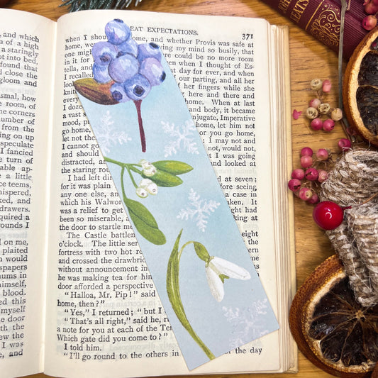 Winter wanderland botanical floral bookmark illustrated with snowdrop, mistletoe and blueberry, cut tit he shape of the blueberries at the top, resting in open pages of a book