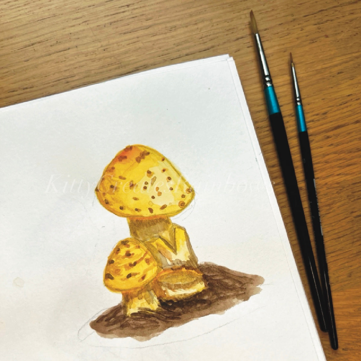 Yellow amanita watercolour original painting by Kat Lovatt on a wooden desk with 2 paintbrushes laying next to the sketchbook