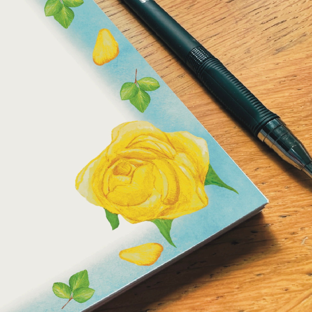 Close up corner of yellow rose illustrated blank notepad with sky blue border decorated with yellow petals and leaves