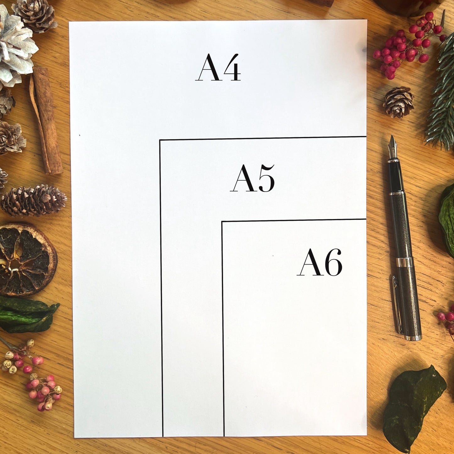 Paper size sample shows an A4 sheet of paper on a table with winter decor and a fountain pen lying next to it.  The A4 has an A5 indicated as half the size of A4 and then an A6 indicated as half the size of A5