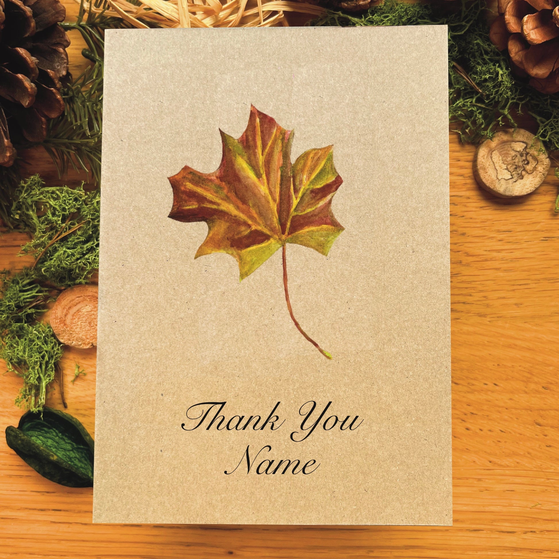 Maple leaf illustrated in watercolour attached to Manila recycled greetings card on a wooden desk, with text script Thank You Name underneath the illustration