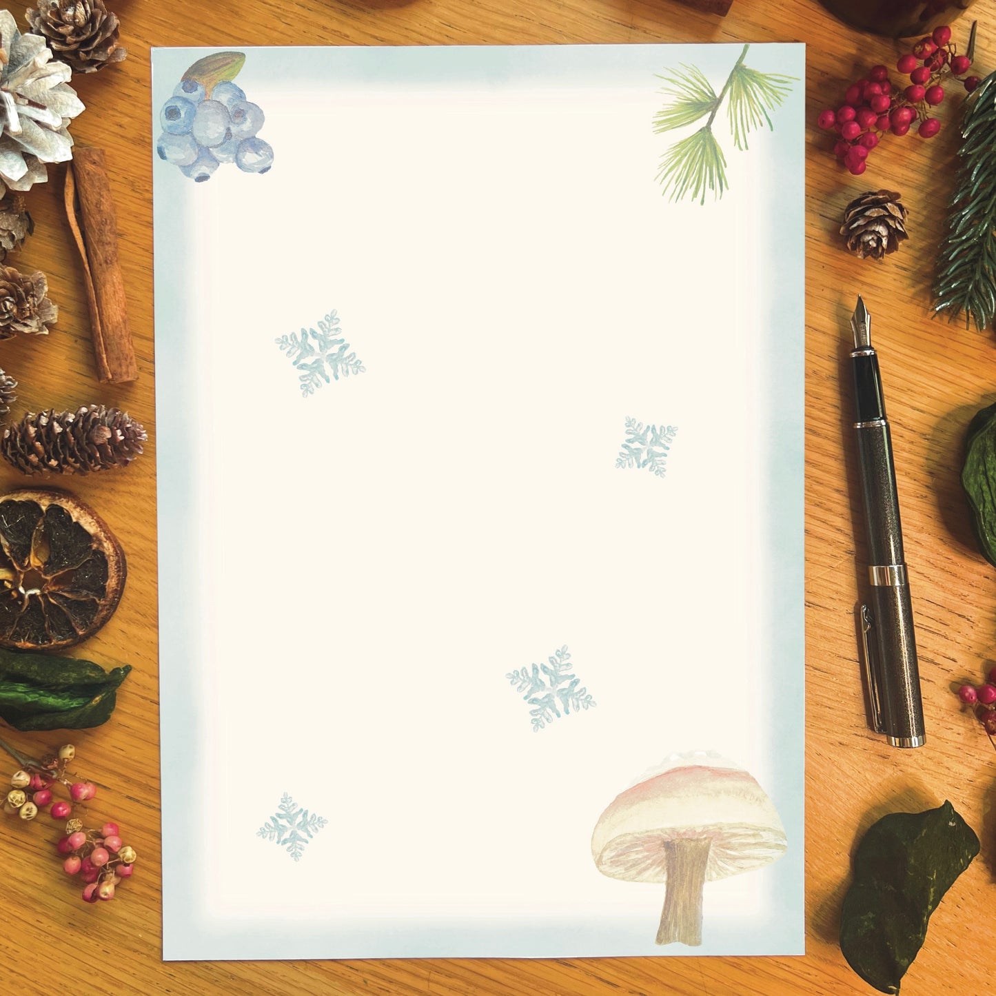Winter wanderland illustrated paper featuring a snowy milkcap mushroom with an ice blue border and snowflakes across the page, on a wooden desk
