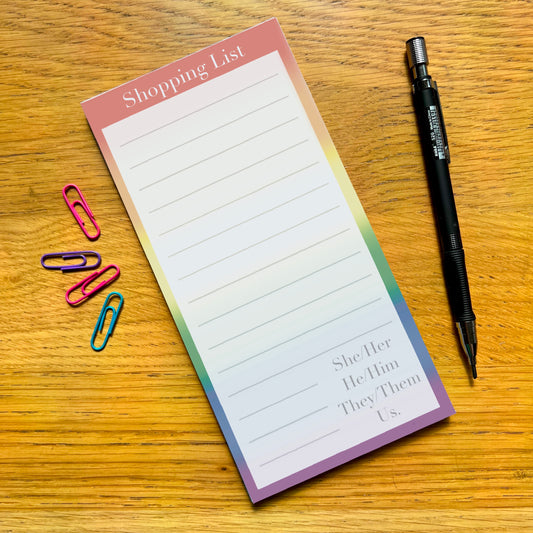 Rainbow shopping list pad with quote She/her He/him They/them Us, on a wooden desk