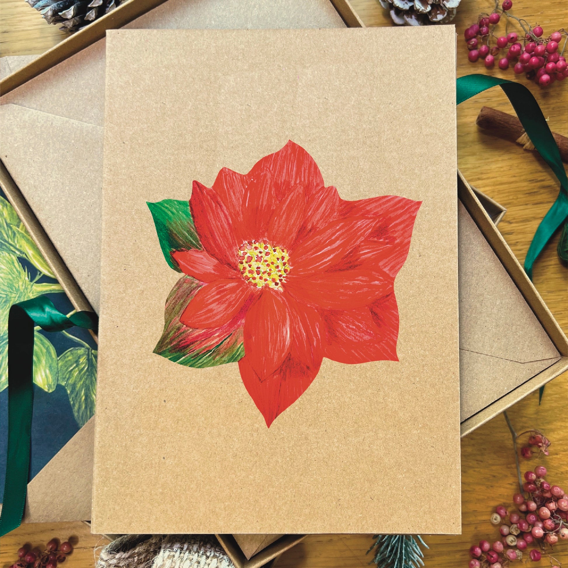 Red poinsettia illustrated in watercolour attached to brown Manila recycled greetings card on a wooden desk
