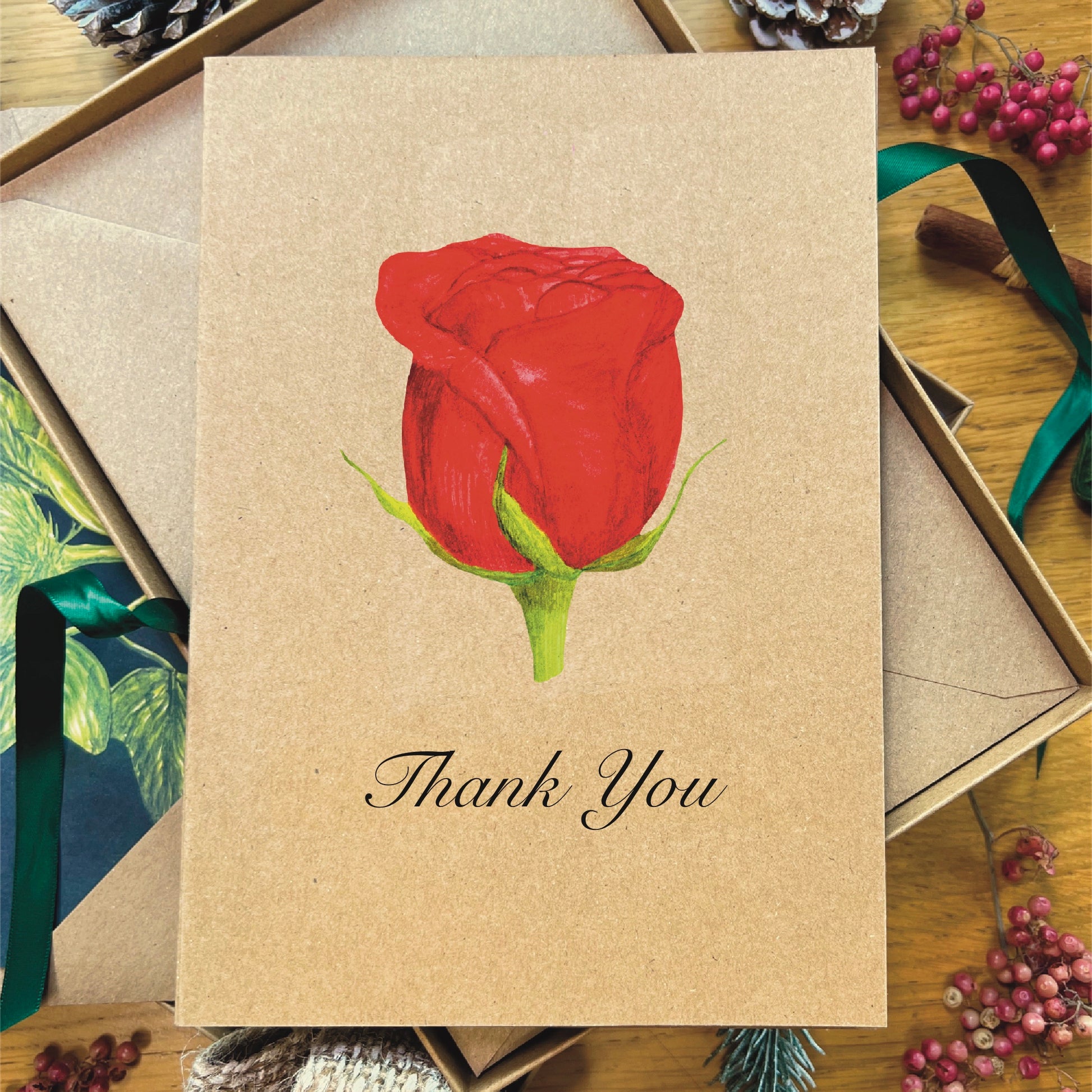 Red hybrid tea rose painted in watercolour attached to a natural recycled blank greetings card, laying in a box with an illustrated inlay envelope with Thank You written in script underneath the rose