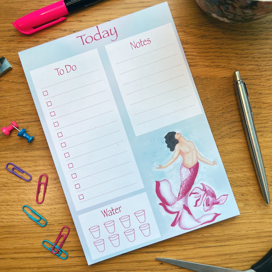 Blossom planner pad resting on a wooden desk decorated and surrounded by pens and other stationery items.  Planner has 3 white box sections, left To Do, right Notes and bottom left Water tracker with illustrated glasses. Blossom mermaid is illustrated bottom right. Blossom has a light and magenta pink tail shaped like petals, with Caucasian skin tone, subtle top surgery scars and mid length black hair flowing. A matching colours pink koi swims next them.