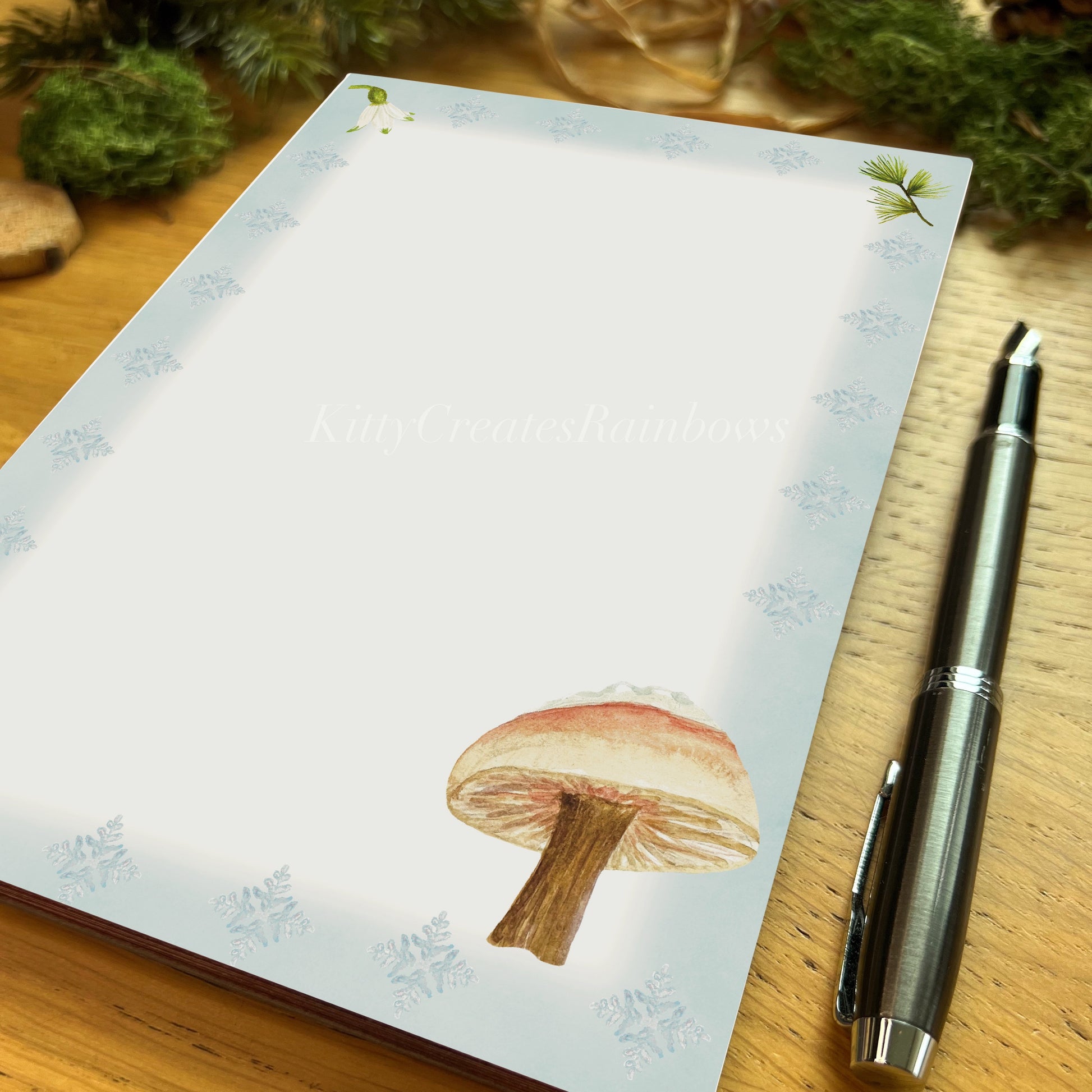 Snow topped milkcap mushroom illustrated notepad page with icy blue border on a wooden desk with a fountain pen