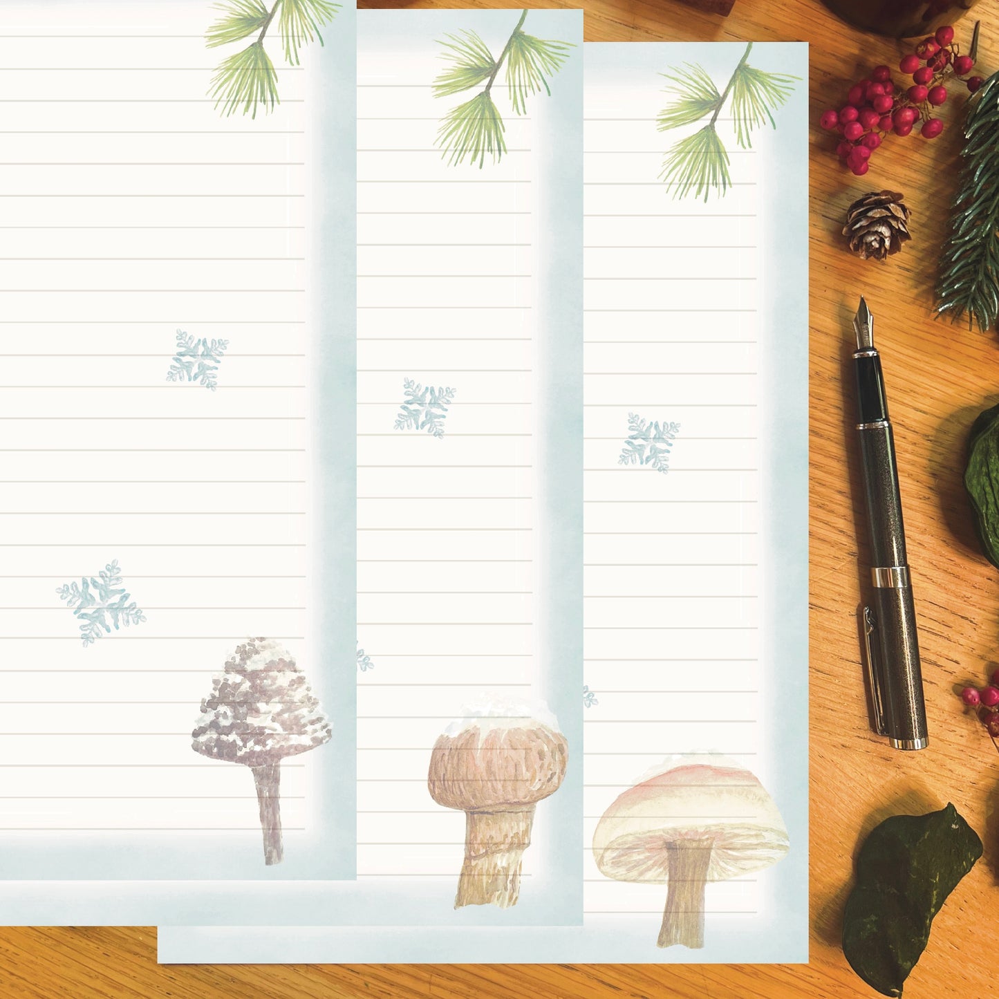 3 Winter wanderland illustrated papers featuring a snow topped mushroom with an ice blue border and snowflakes across the page, on a wooden desk