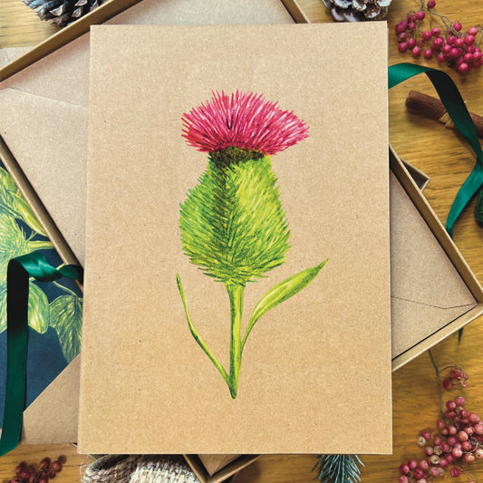 Spear thistle illustrated in watercolour attached to brown Manila recycled greetings card on a wooden desk