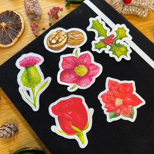 Winters bloom die cut stickers including red rose, hellebore, poinsettia, holly, thistle and walnut, on a black journal