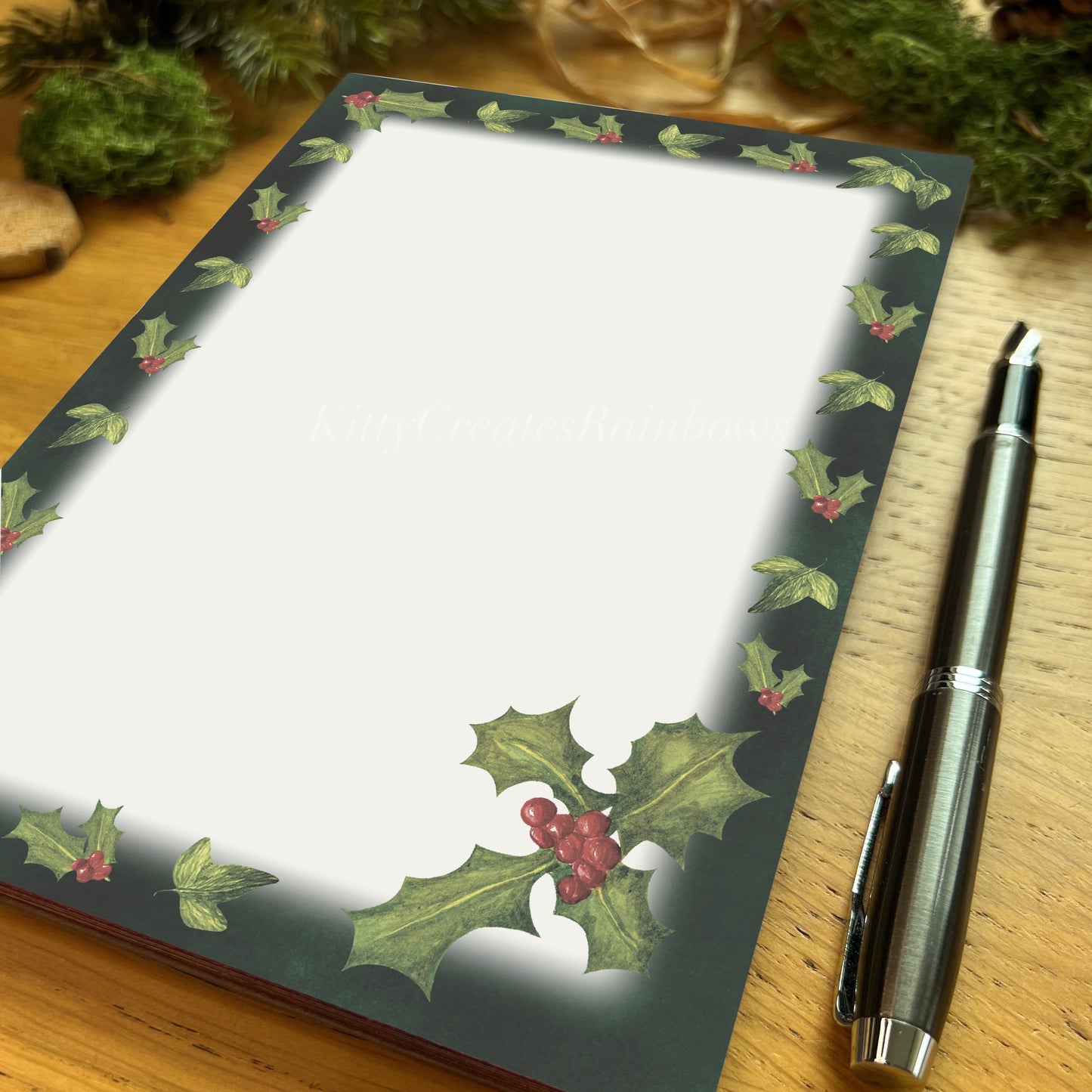 Winters bloom Holly sprig with red berries Winters bloom red rose illustrated notepad with dark evergreen border, on a wooden table with fountain pen
