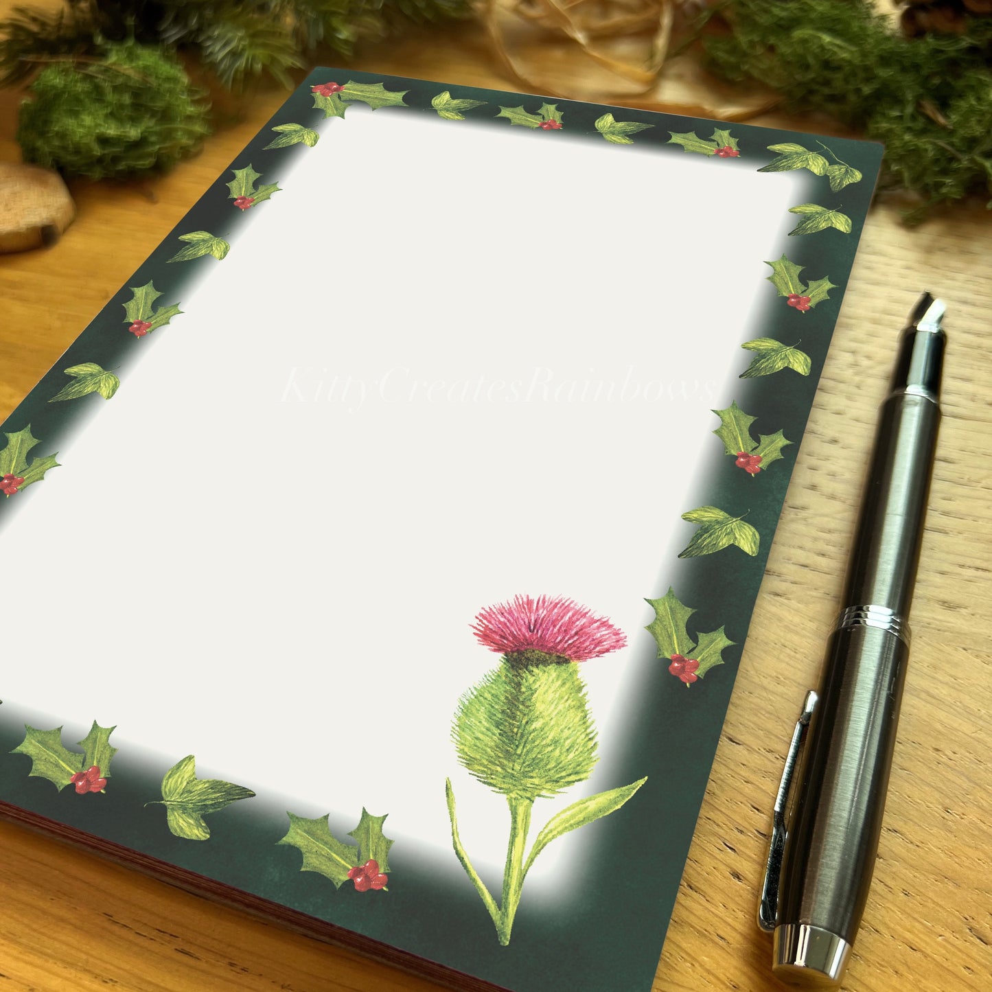 Winters bloom thistle illustrated notepad with dark evergreen border, on a wooden table with fountain pen