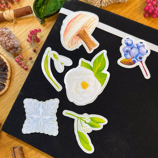 Winter wanderland die cut stickers of white rose, mistletoe, snowdrop and snow topped wild mushrooms laid on a black journal